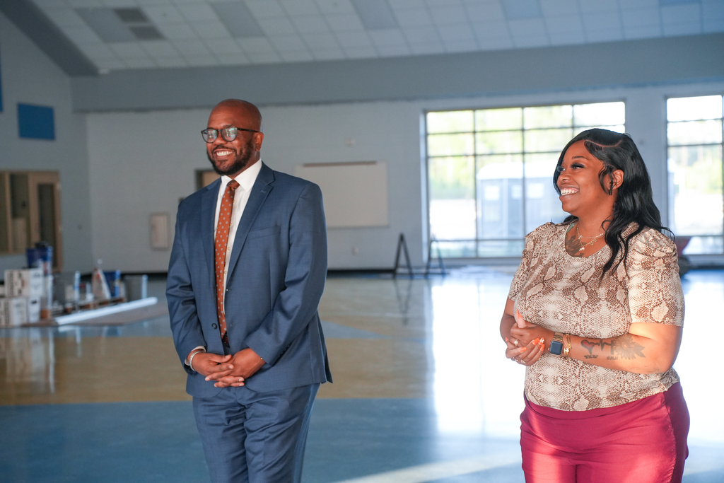 A man in a suit and a woman with tattoos in a large, empty gym.