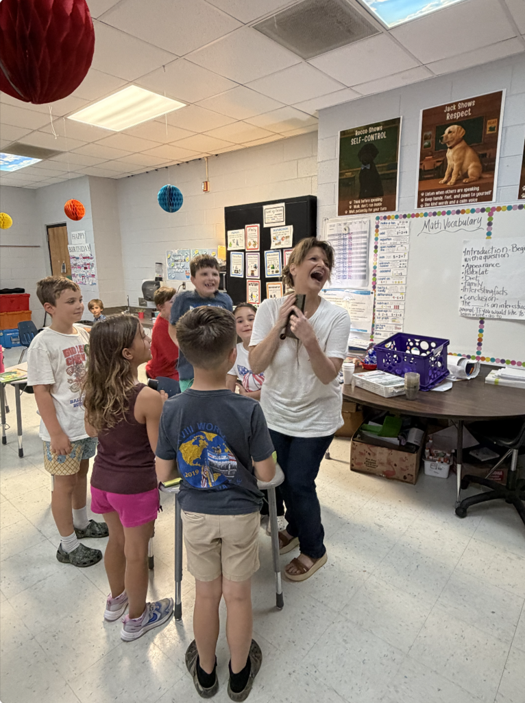 A teacher with a microphone speaks to a group of children standing in a classroom.