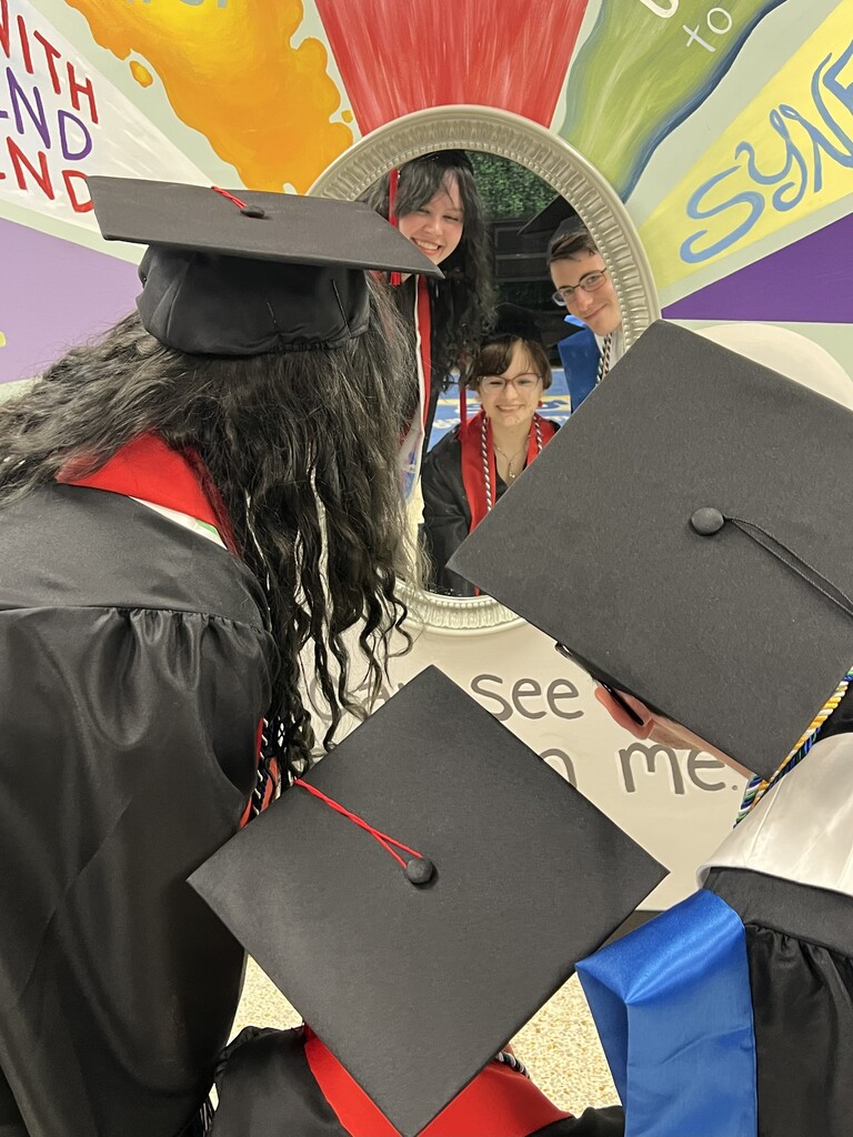 Three people in black graduation gowns look at a mirror. The mirror reflects a woman and man.