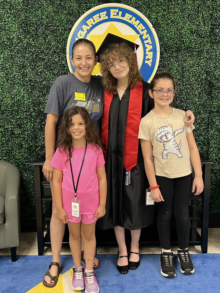 Four children pose in front of a green backdrop with a school logo. The two girls wear pink and tan shirts. A graduate wears a black gown with a red stole.