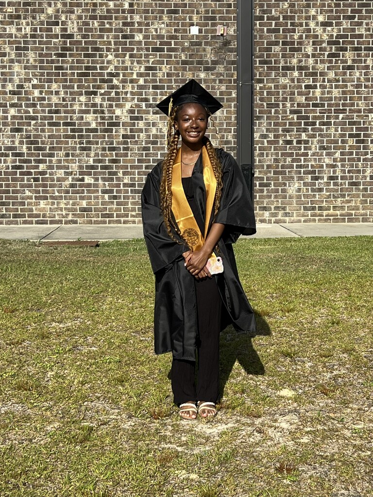 A person in a graduation gown and cap stands on a grassy area in front of a brick wall.