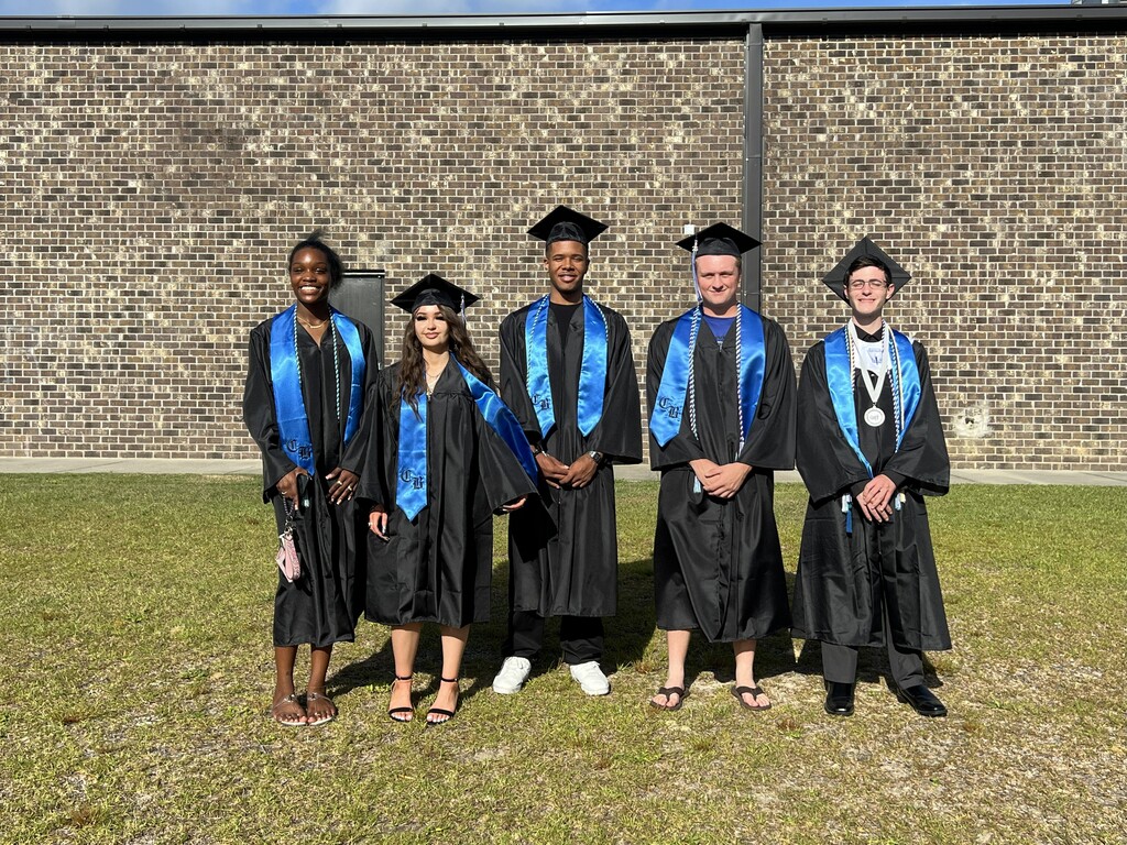 Five people in black and blue graduation gowns stand on grass in front of a brick wall.