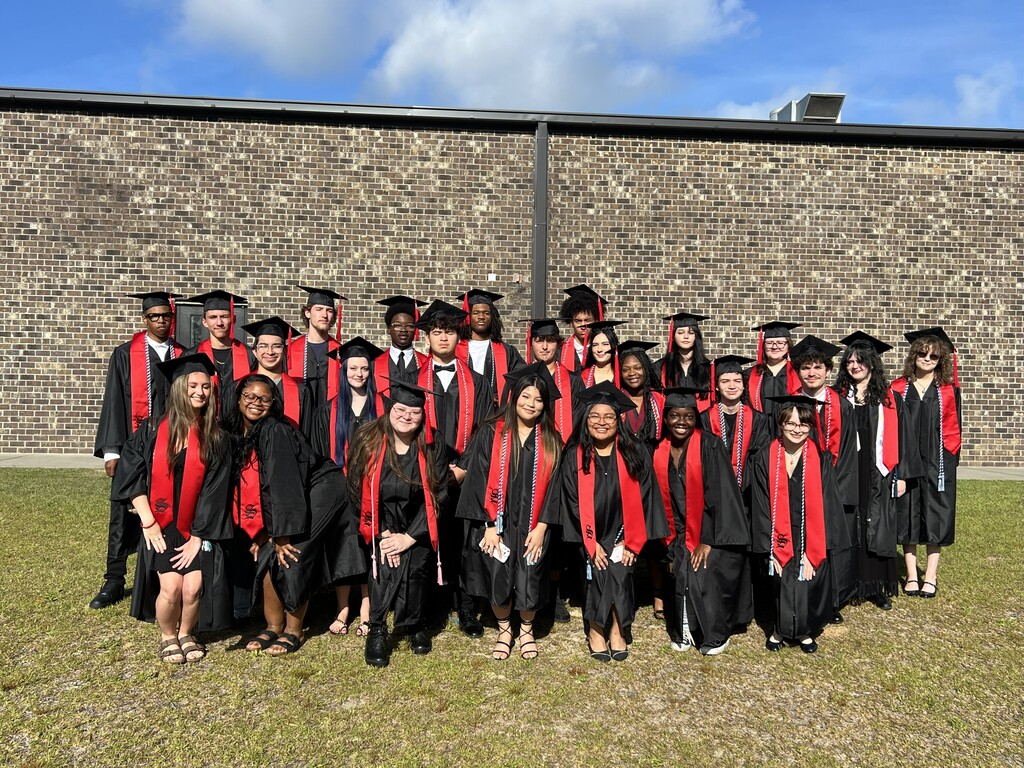 A group of graduates in black and red caps and gowns stands in front of a stone wall.