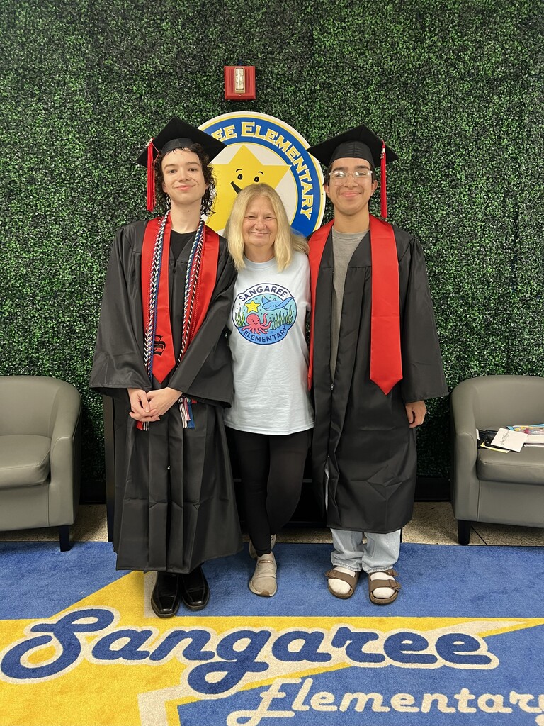 Three people in graduation attire stand against a green wall with a school logo, one in a cap and gown, another in a cap, gown, and red stole, and a third in a white shirt.