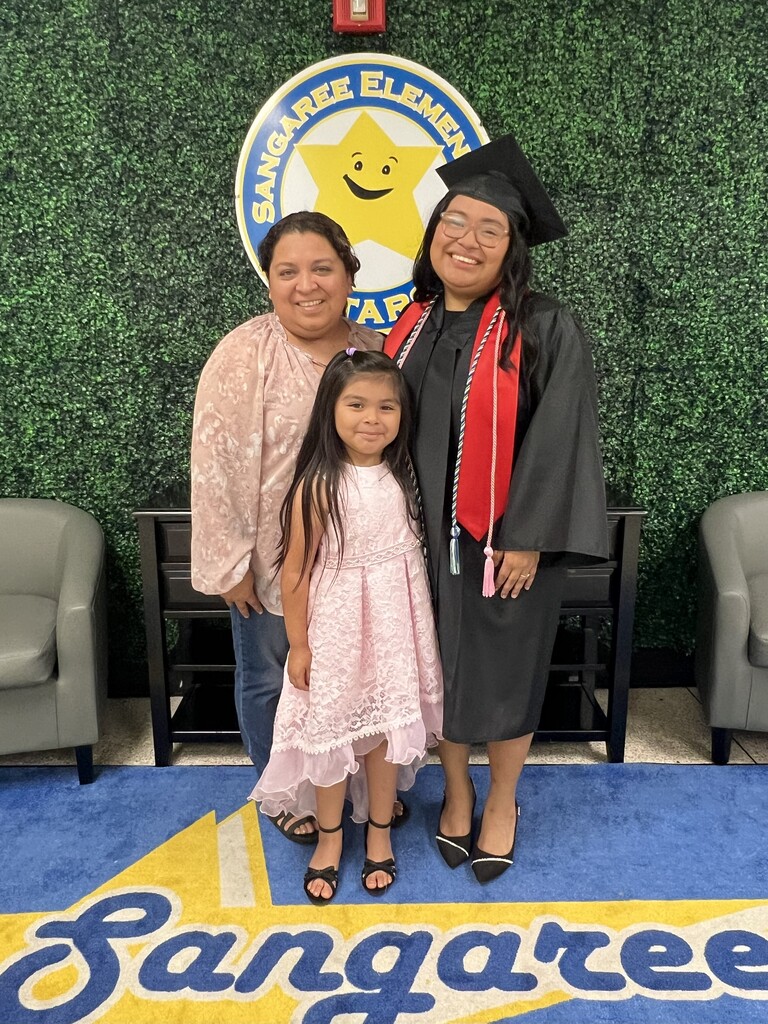 Three individuals in front of a wall with the words "Sangaree Elementary." A woman and a girl wear pink, another woman in a graduation gown.