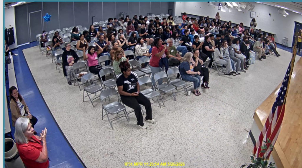 A ceremony with people sitting in chairs, with one person standing and clapping in front of a flag.