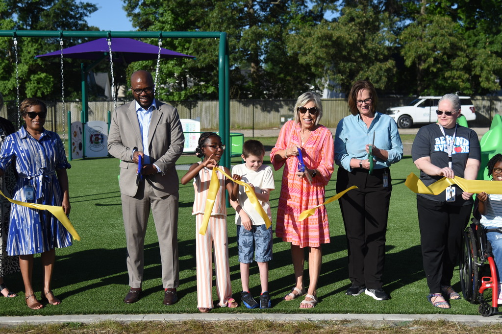A group of people stand on artificial grass with a ribbon, likely celebrating an opening. A child is in a wheelchair.