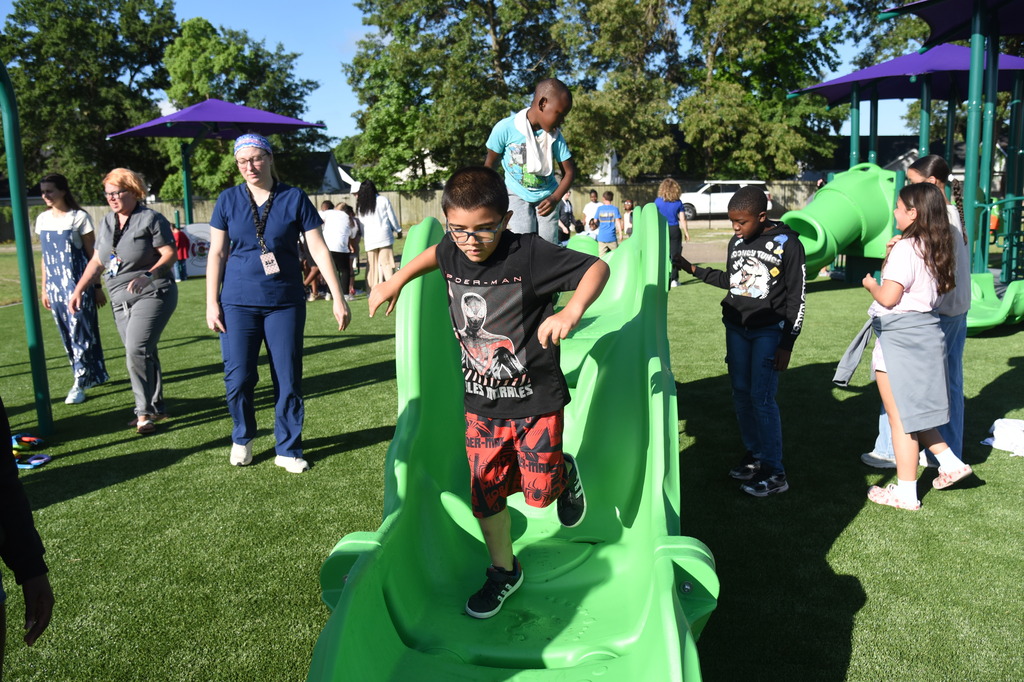 Playground with artificial turf. A boy in glasses slides down a green slide. Several children, adults, and trees in the background.