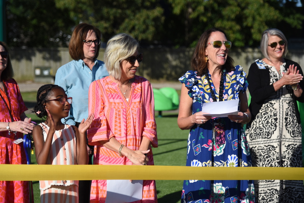 People in colorful attire gather on a field, with a woman holding a paper and speaking into a microphone.