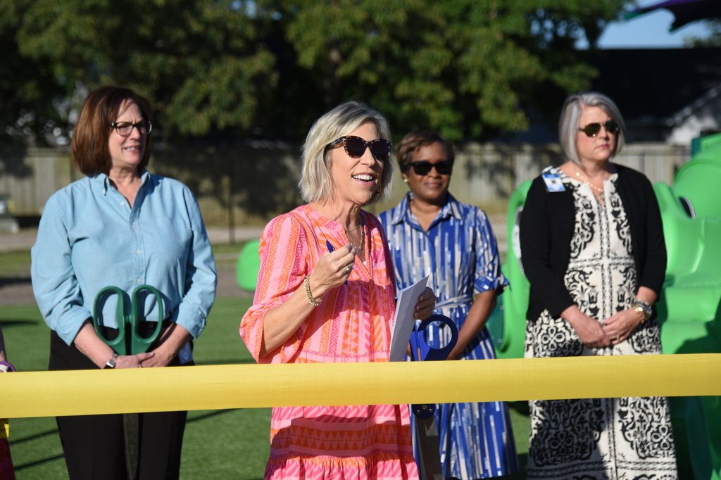 Four women stand in front of a yellow ribbon. One holds a paper, another a pen. Green structure and trees behind.