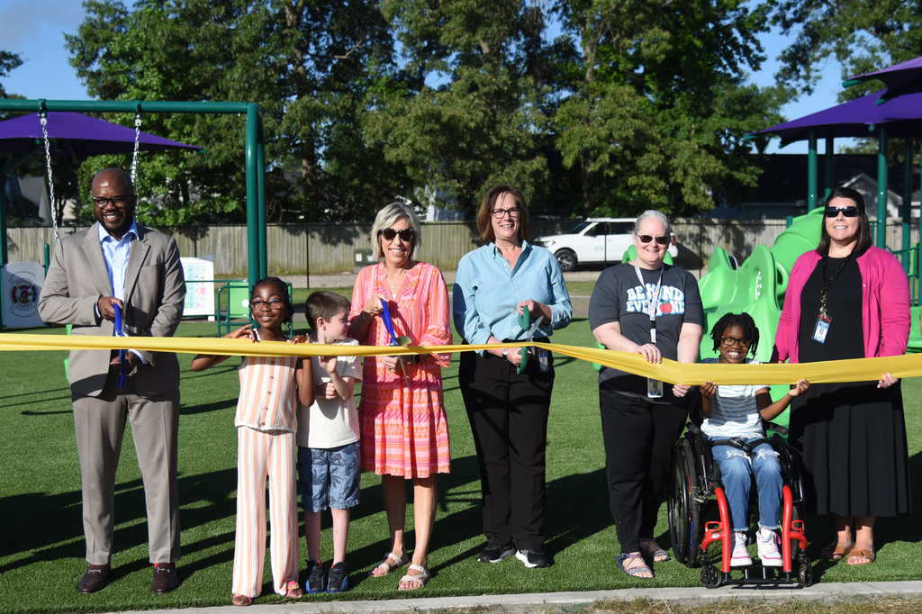 People gather on a green lawn, holding a ribbon. Some are standing, others in wheelchairs. A few trees and play equipment are in the background.