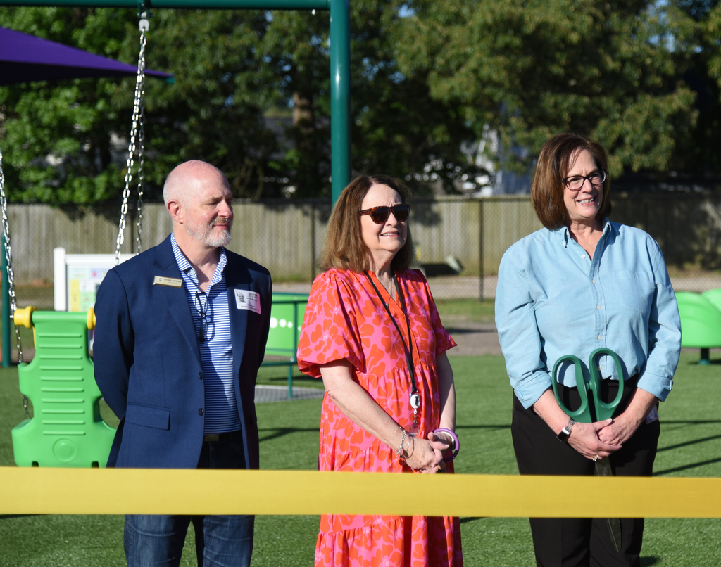 Three people stand before a ribbon. One holds scissors. A playground with swings and a fence is behind them.