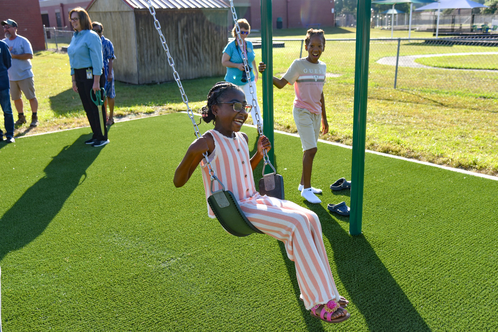 A child sits on a swing while others stand nearby on a green artificial turf surface.