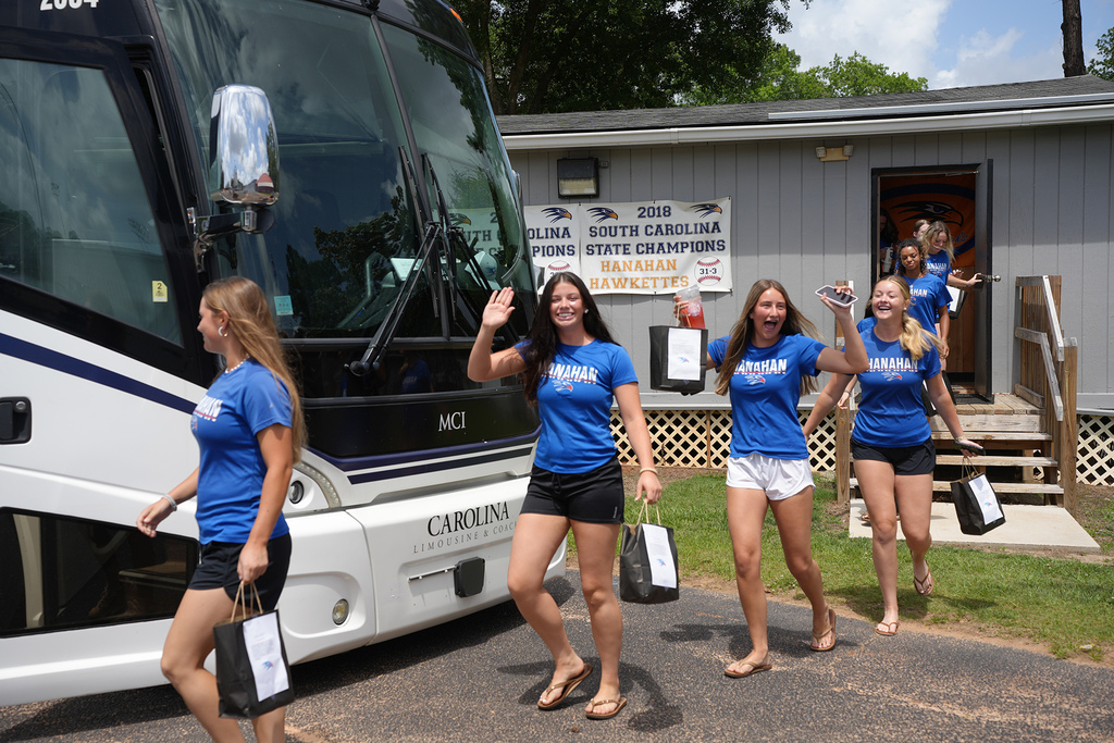 Five women in matching blue shirts and shorts are walking in front of a bus. They hold bags in their hands.