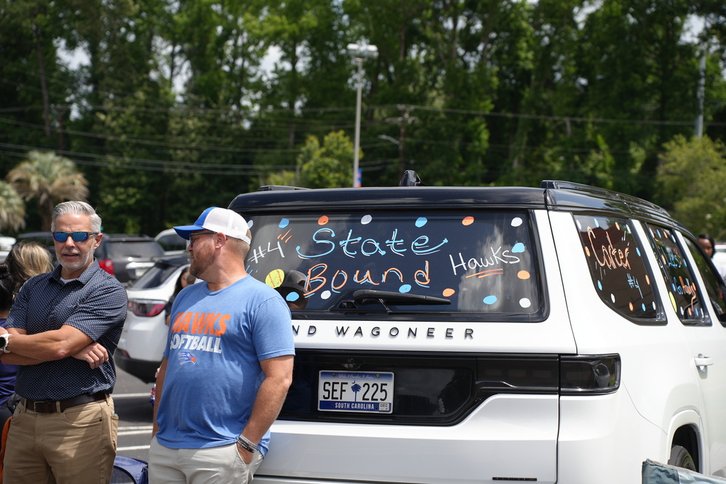 Two men, one in a blue shirt and the other in a white hat, stand by a white car. The car's back window has colorful decorations. Trees and parked cars are in the background.