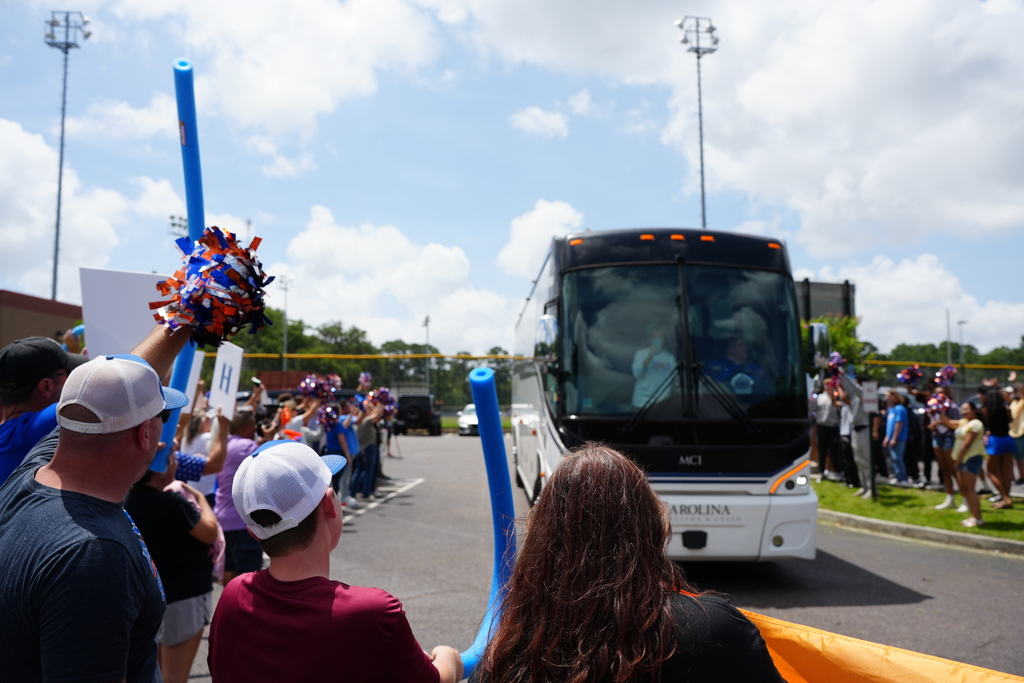 People gather by a road, cheering as a bus passes by. One holds blue pompoms, another wears a white cap.
