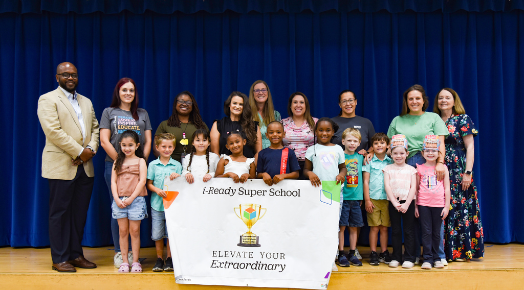 A group of people from Westview Primary, including adults and children, stand behind a banner with a trophy. They are on a stage with a blue curtain backdrop.