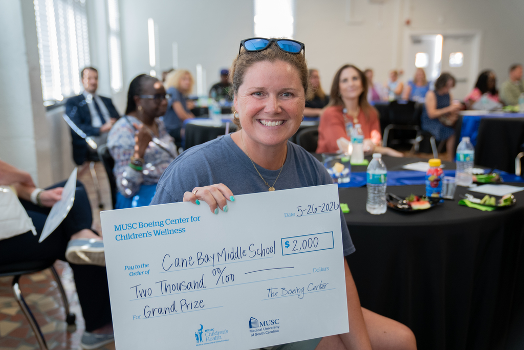 A smiling person holds a check for $2,000, labeled "Grand Prize", standing in a room with tables and chairs.