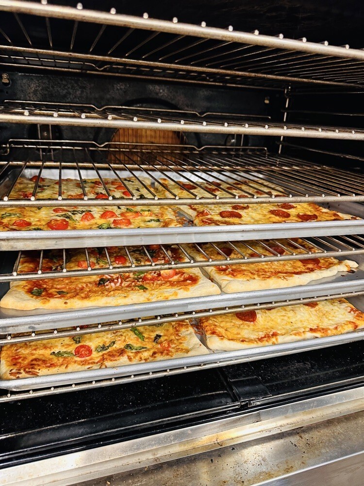 Four pizzas with various toppings are arranged on three racks in an oven. The oven has a black interior and stainless steel racks.