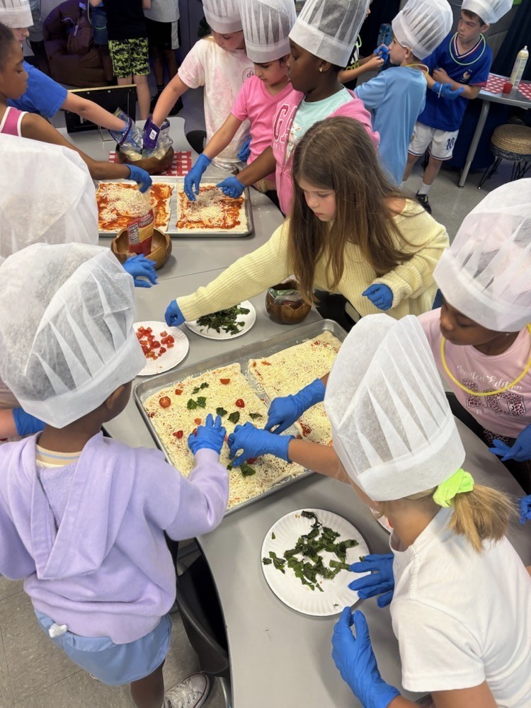 A group of children in hairnets and gloves are making pizza on trays at a table.