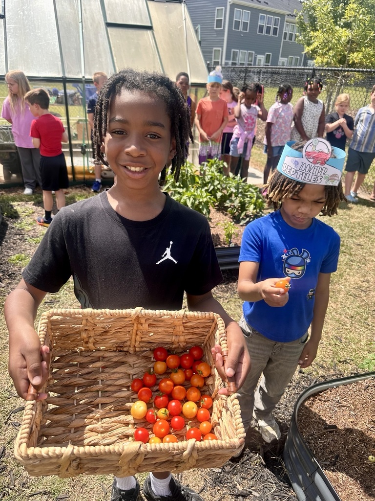 Two children stand outside with a basket of tomatoes. Behind them are a crowd of people, a garden, and a greenhouse.