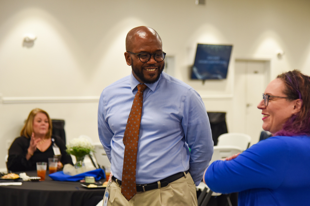 A man in a light blue shirt and brown tie converses with a woman in a blue shirt, both wearing glasses. Behind them, a woman sits at a table with drinks and flowers.