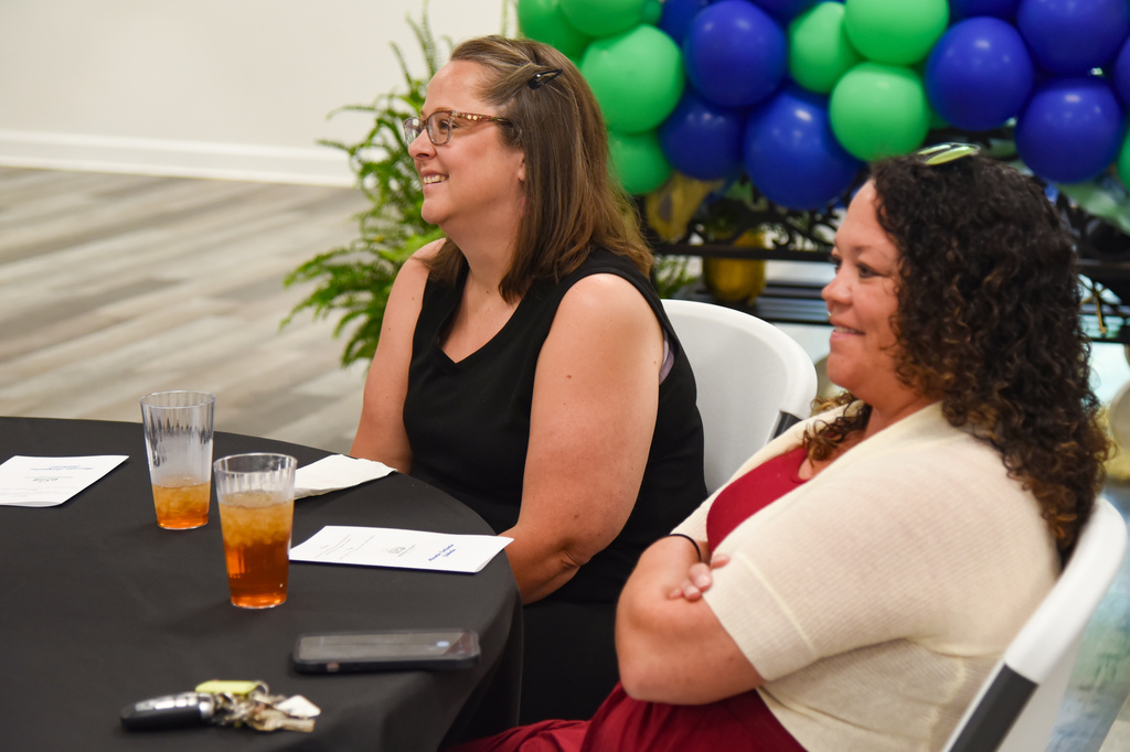 Two women sit at a table, one with glasses and a black top, another with curly hair and a beige cardigan.