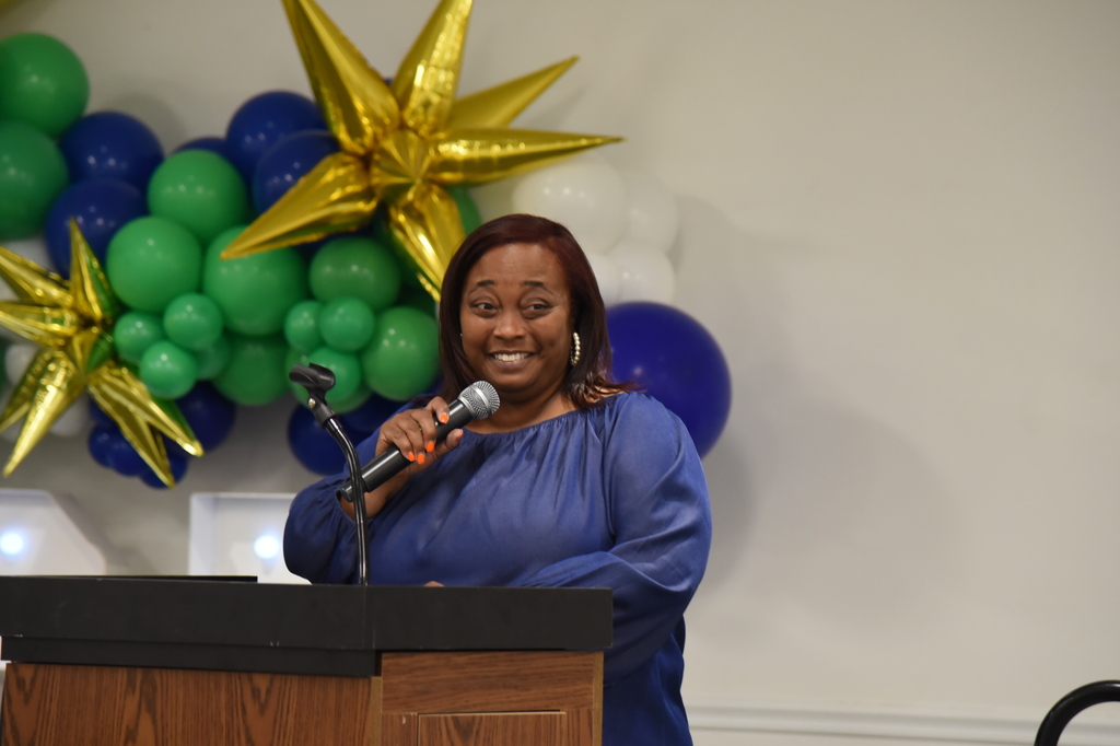 A woman in a blue top speaks into a microphone at a podium, with a white wall and colorful balloons in the background.
