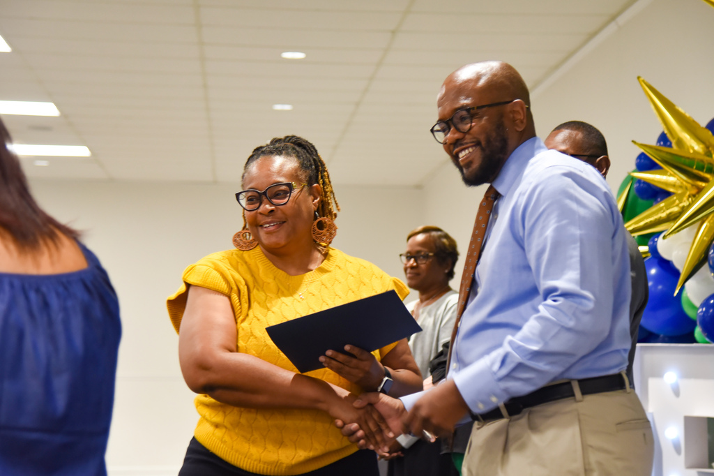 People in a room, one woman hands a folder to a man, both wearing glasses, they shake hands.