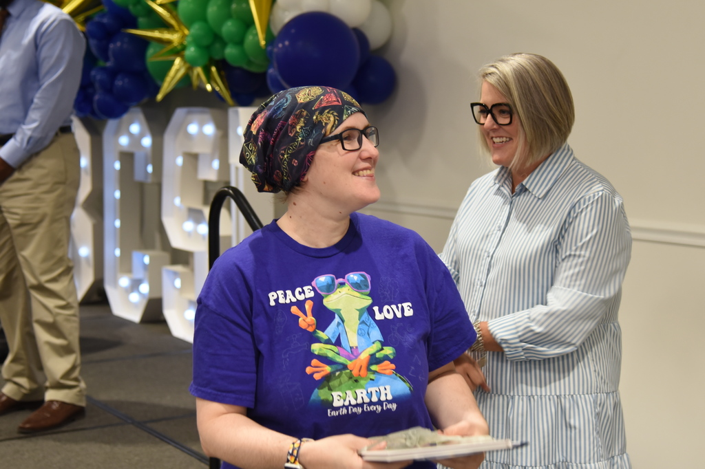 Two women smiling, one wearing glasses and a headband, standing in front of a large number sign.