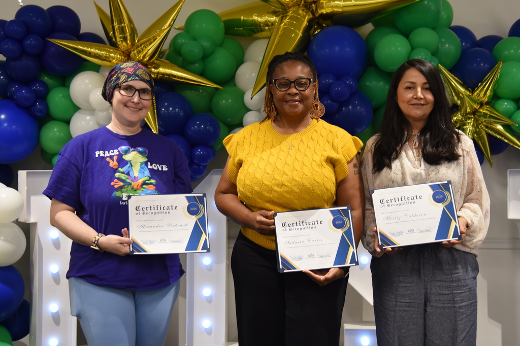 Three women stand holding certificates in front of a wall of balloons and lights.