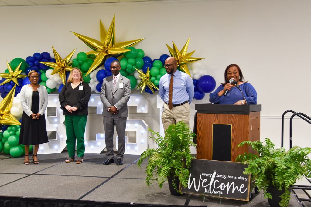A group of six people on stage, a woman at a podium speaking into a microphone, others standing behind. The stage is decorated with balloons and plants.