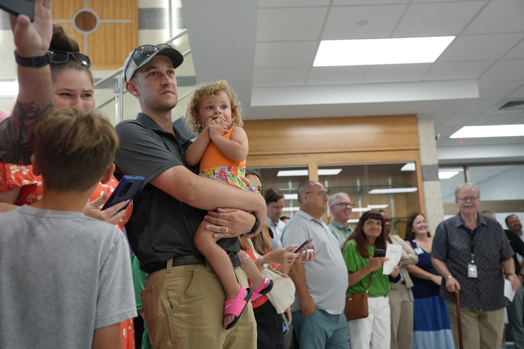 Group of people, including a man holding a child, standing in a hallway with glass walls.