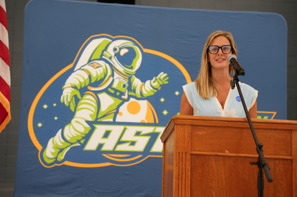 Woman at podium with microphone and blue backdrop featuring a logo of a jumping astronaut in a spacesuit.