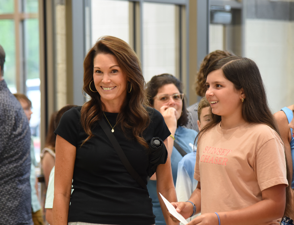 A woman with dark hair stands with a girl in front of a building. Both are smiling.
