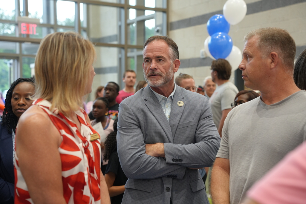 Three adults converse in a large room with a crowd of people and balloons in the background.