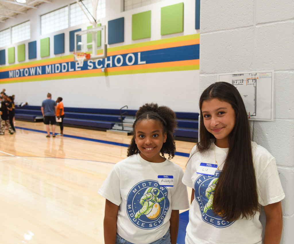 Two girls in white shirts stand near a wall in a gymnasium, smiling. Behind them, a basketball court and a banner reading Midtown Middle School.