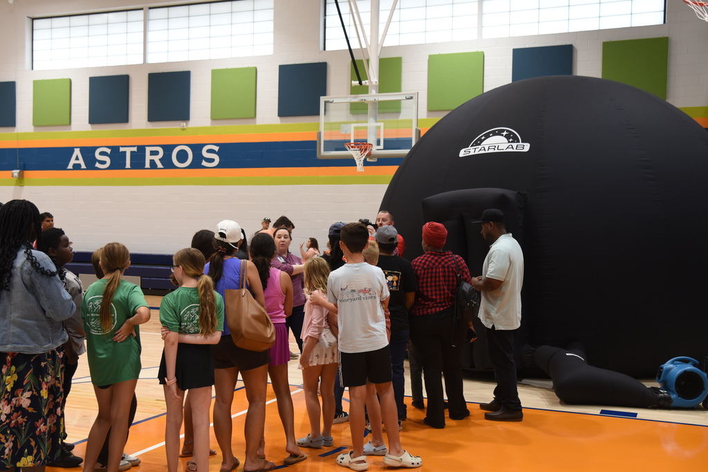 A group of children and adults gather near a black inflatable dome in a gymnasium.