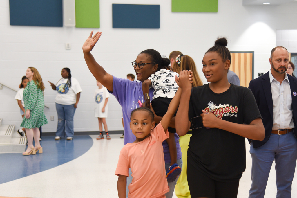 Indoors, several people stand, some with arms raised. A woman waves with a young child. Another holds a toddler, while others observe in the background.