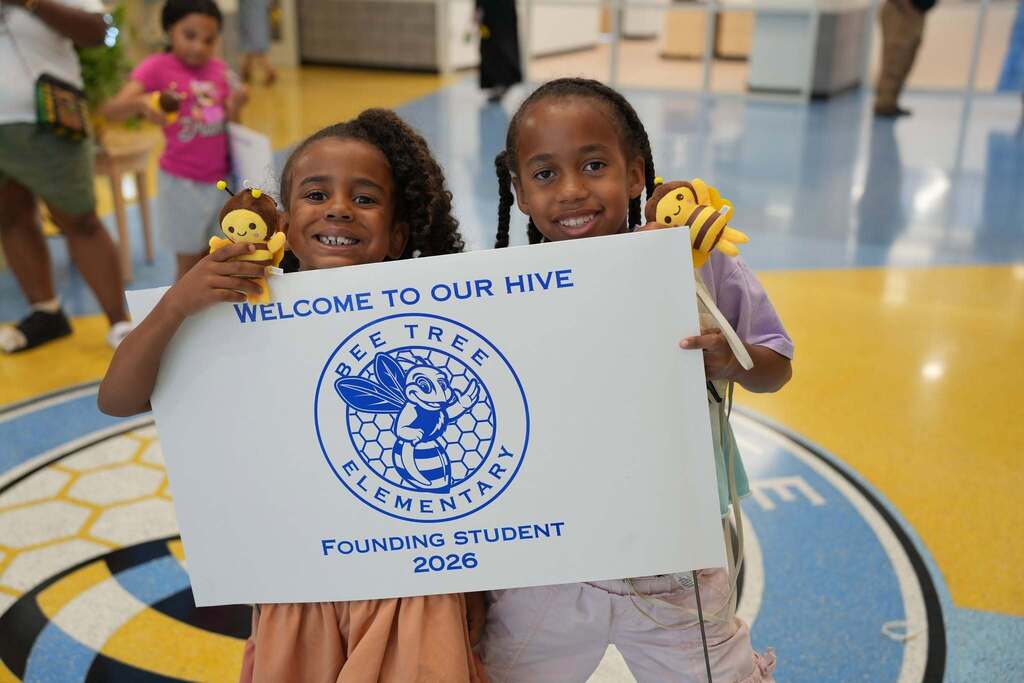 Two children hold a sign that reads "Welcome to our hive" at a school event. The sign features a bee logo and "Founding Student 2016" text.