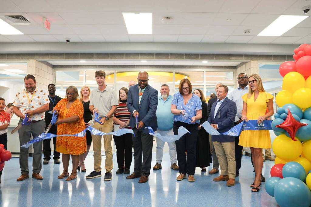 Group of adults at a ribbon-cutting event, standing in front of balloons. Brightly lit room with white walls and a tiled floor.