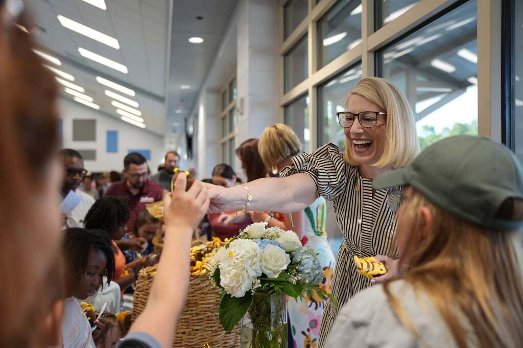 Woman with glasses and striped shirt hands basket of flowers to another person. They are surrounded by others.