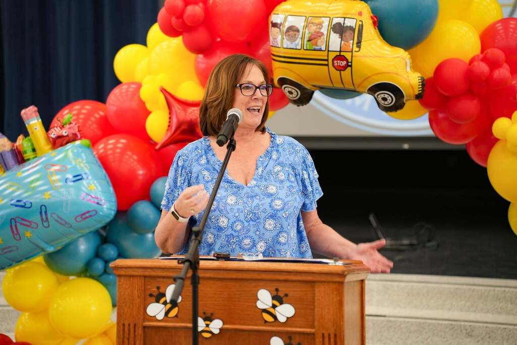 A woman stands at a podium with a microphone, addressing an audience. Behind her, a colorful balloon arch decorates the background.