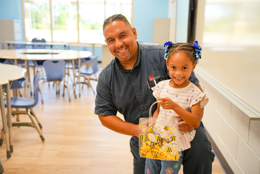 A man and a child smile while standing in a room with a table, chairs, and a window.