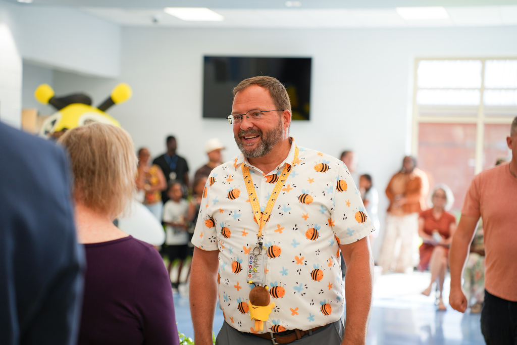 A man wearing a shirt with a bee pattern and an ID lanyard stands in a room with others.