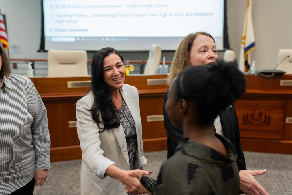 A group of women in business attire are shaking hands in a conference room. Behind them is a projection screen.