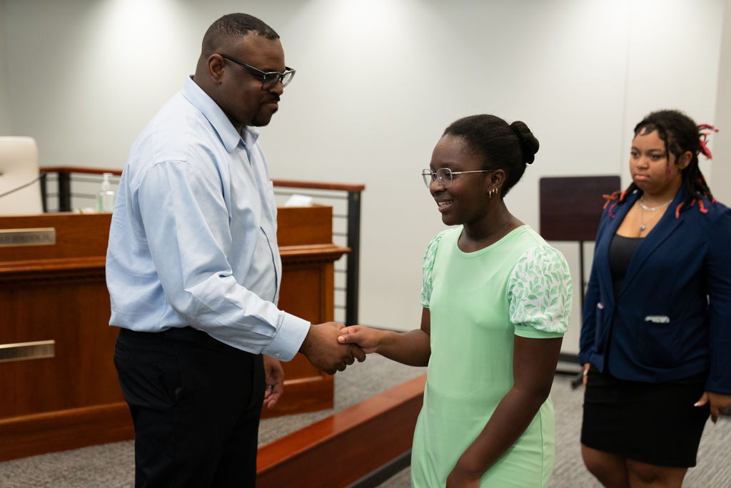 A man in a blue shirt shakes hands with a woman in a green dress in a courtroom.