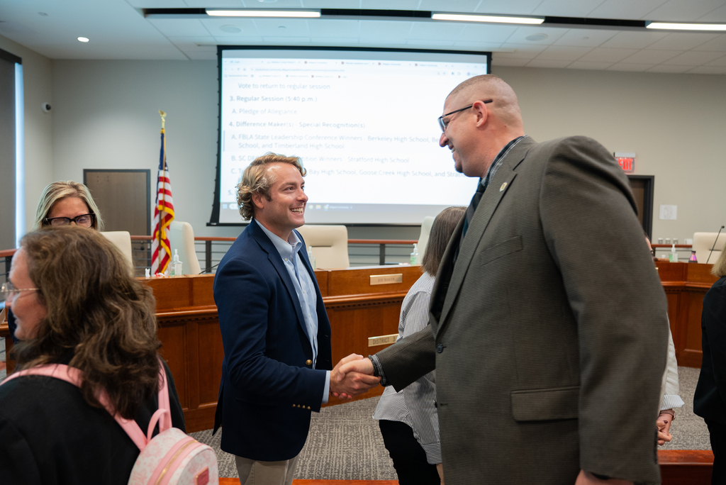 Two men shake hands in an office; one wears a navy blazer and the other a gray suit. Behind them, others are present in a conference room.