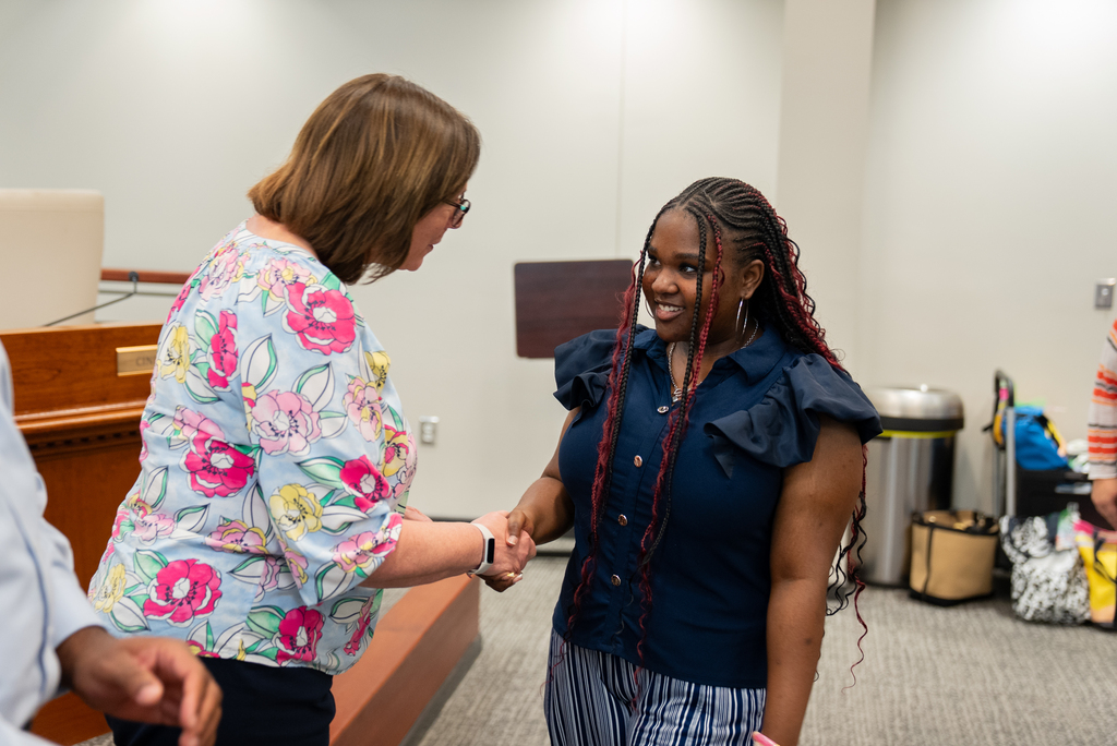 Two women shake hands in a conference room with a trash bin and bags in the background.