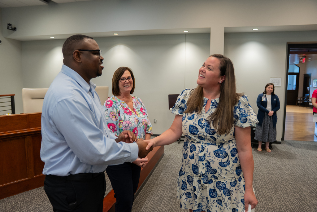 Two people shake hands in a room. One wears a blue dress and the other a blue shirt. Two women stand nearby.